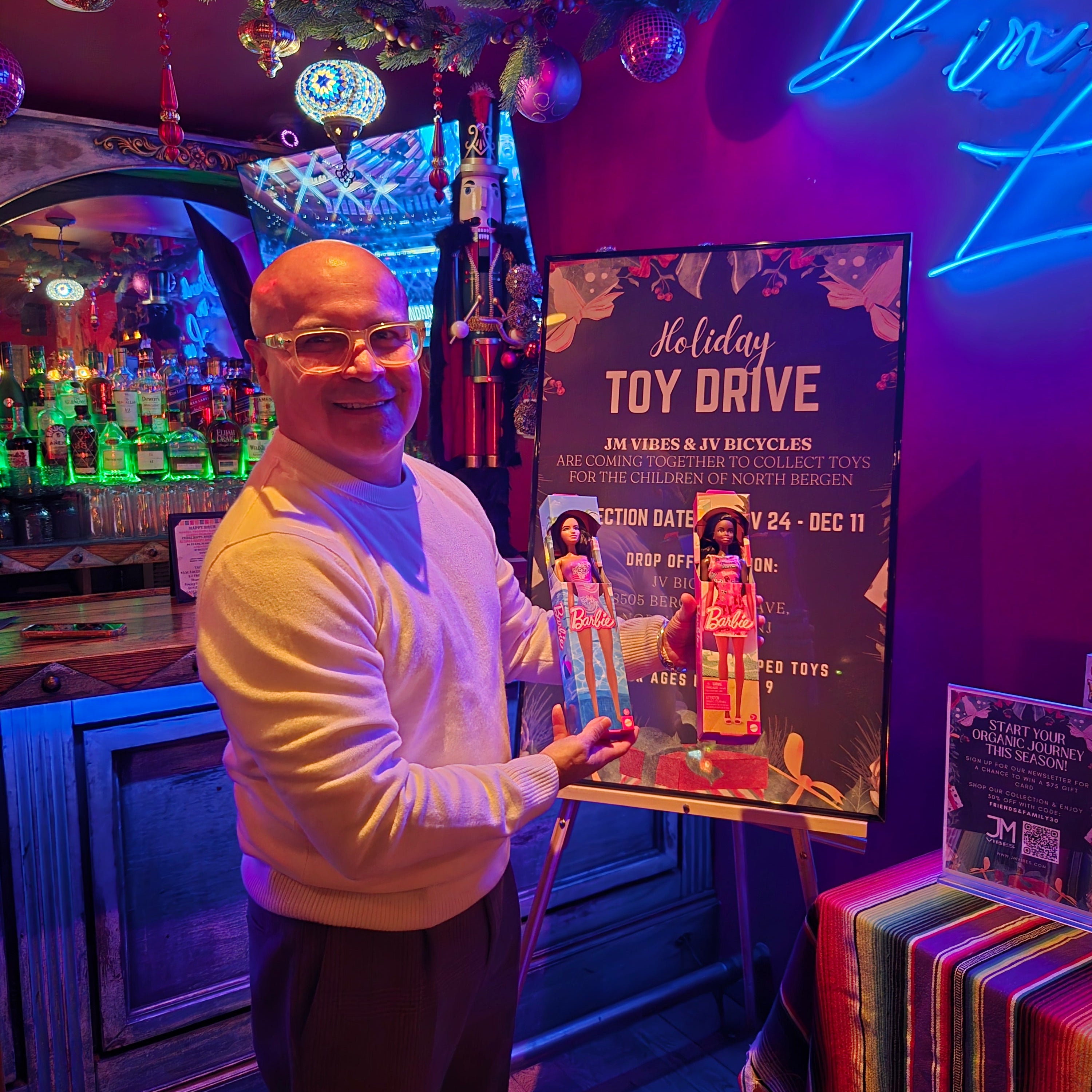 A man in a white long-sleeve shirt and dark trousers stands in a dimly lit bar or club environment with colorful neon lighting, a framed poster, and shelves in the background.