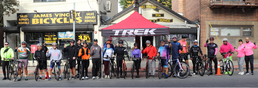 A group of cyclists posing outdoors in front of a brick building with a storefront sign reading James Vincent Bicycles and a red Trek canopy in the background.