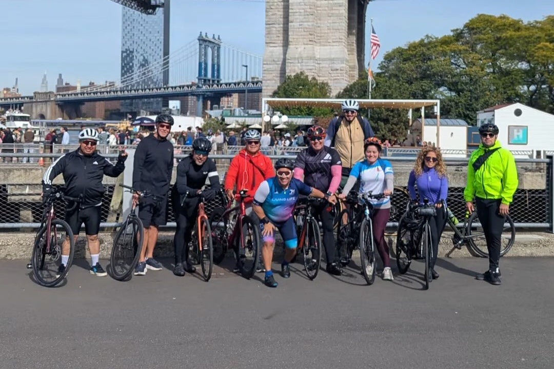 A group of cyclists posing with their bikes on a city street in front of a large suspension bridge under a blue sky.